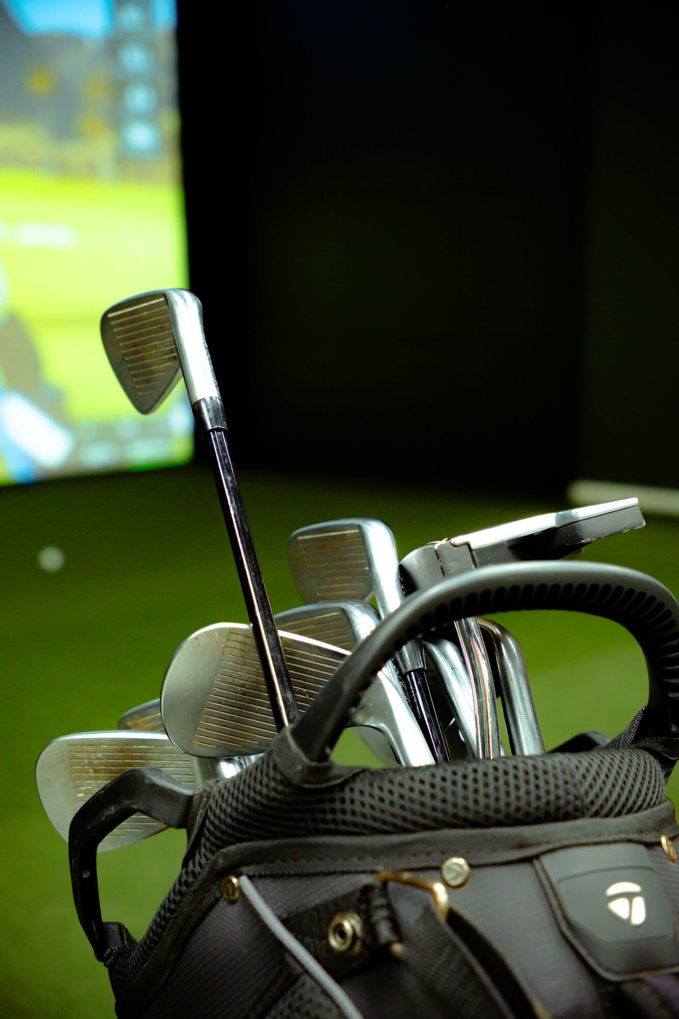 Irons in a golf bag next to a lit simulator screen